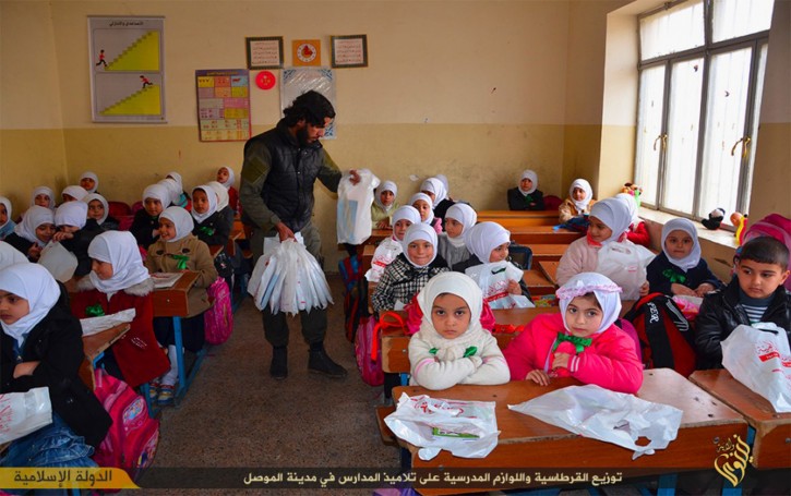 In this photo released on Jan. 11, 2015, by a militant website, which has been verified and is consistent with other AP reporting, an Islamic State militant, center, holds plastic bags full of stationery and other gifts as he distributes them to Iraqi young students, at a school classroom, in Mosul, northern Iraq. Across its territory, IS now runs them, and some parents in IS-run areas take their children out of schools to avoid indoctrination. (Militant website via AP)